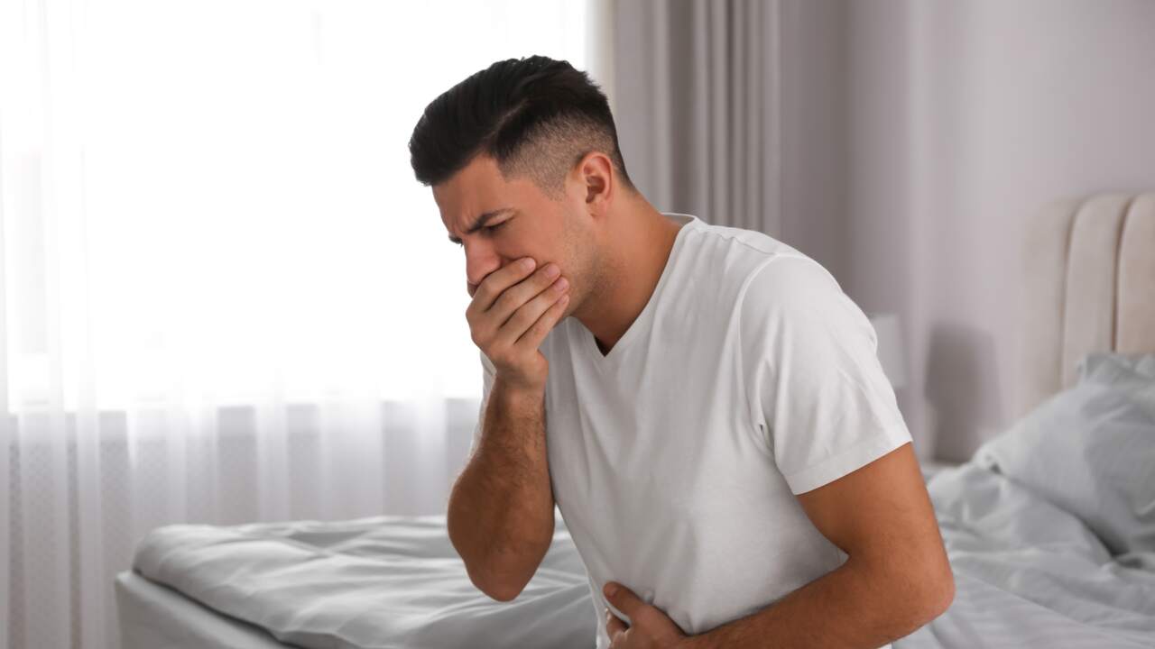 Ein Mann sitzt mit einer Lebensmittelvergiftung auf dem bett und hält sich mit einer Hand den Bauch und mit der anderen den Mund